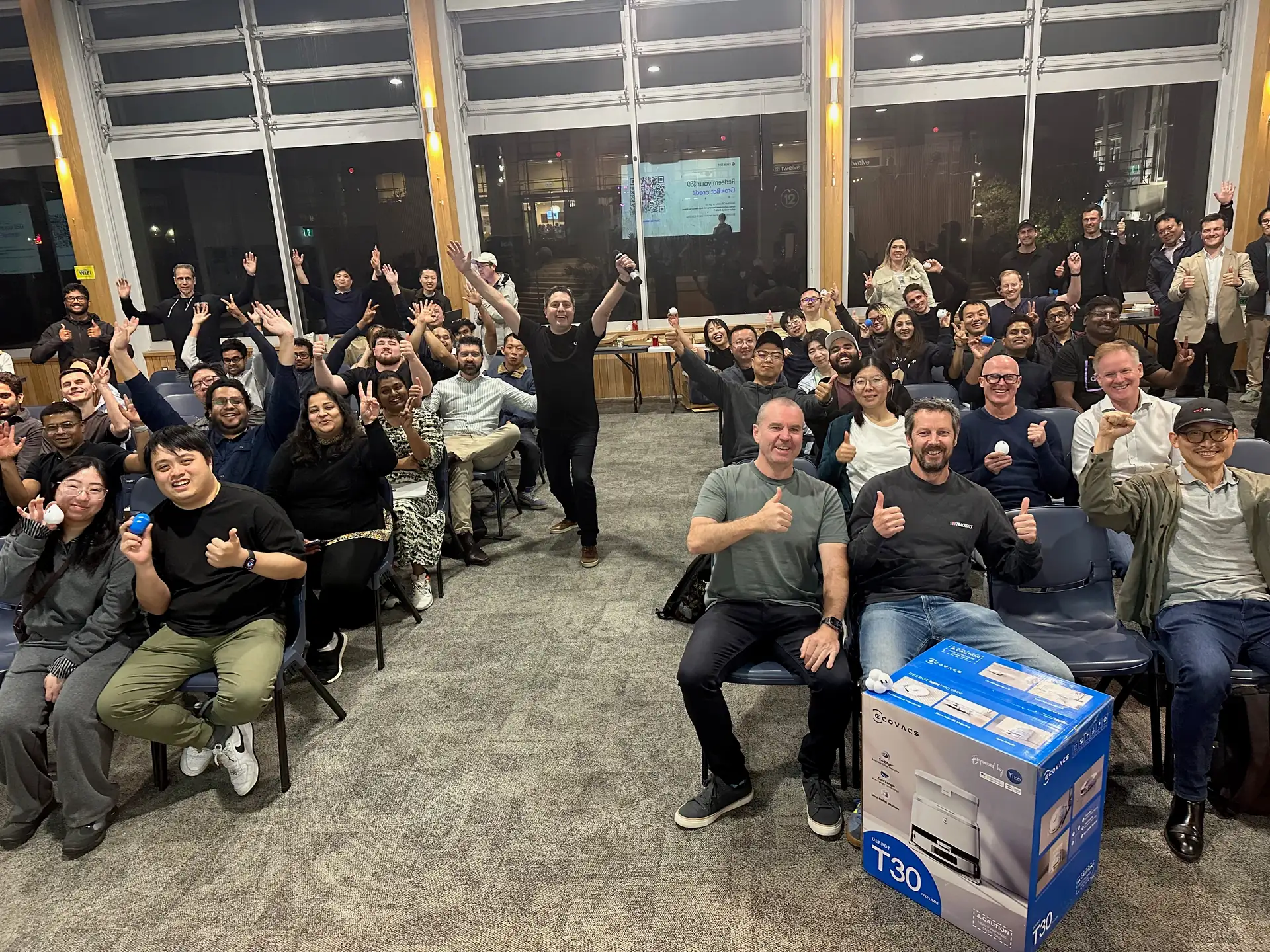 Attendees cheering with hands raised in the end-of-night group photo, with the robot vacuum prize box in front