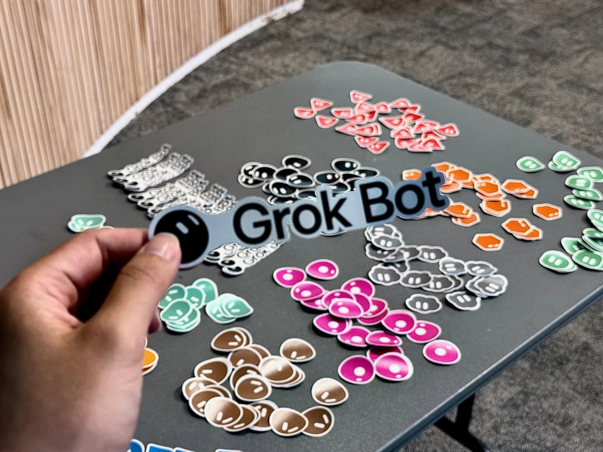 Grok Bot sticker held up over a table of community stickers
