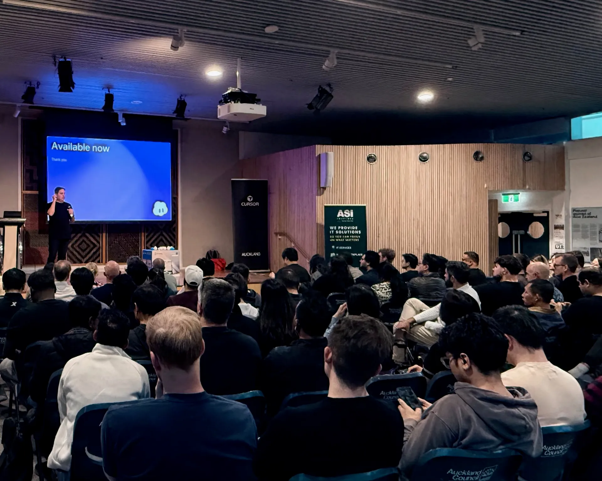 Packed hall seen from the back, with the ASI Solutions banner beside the stage
