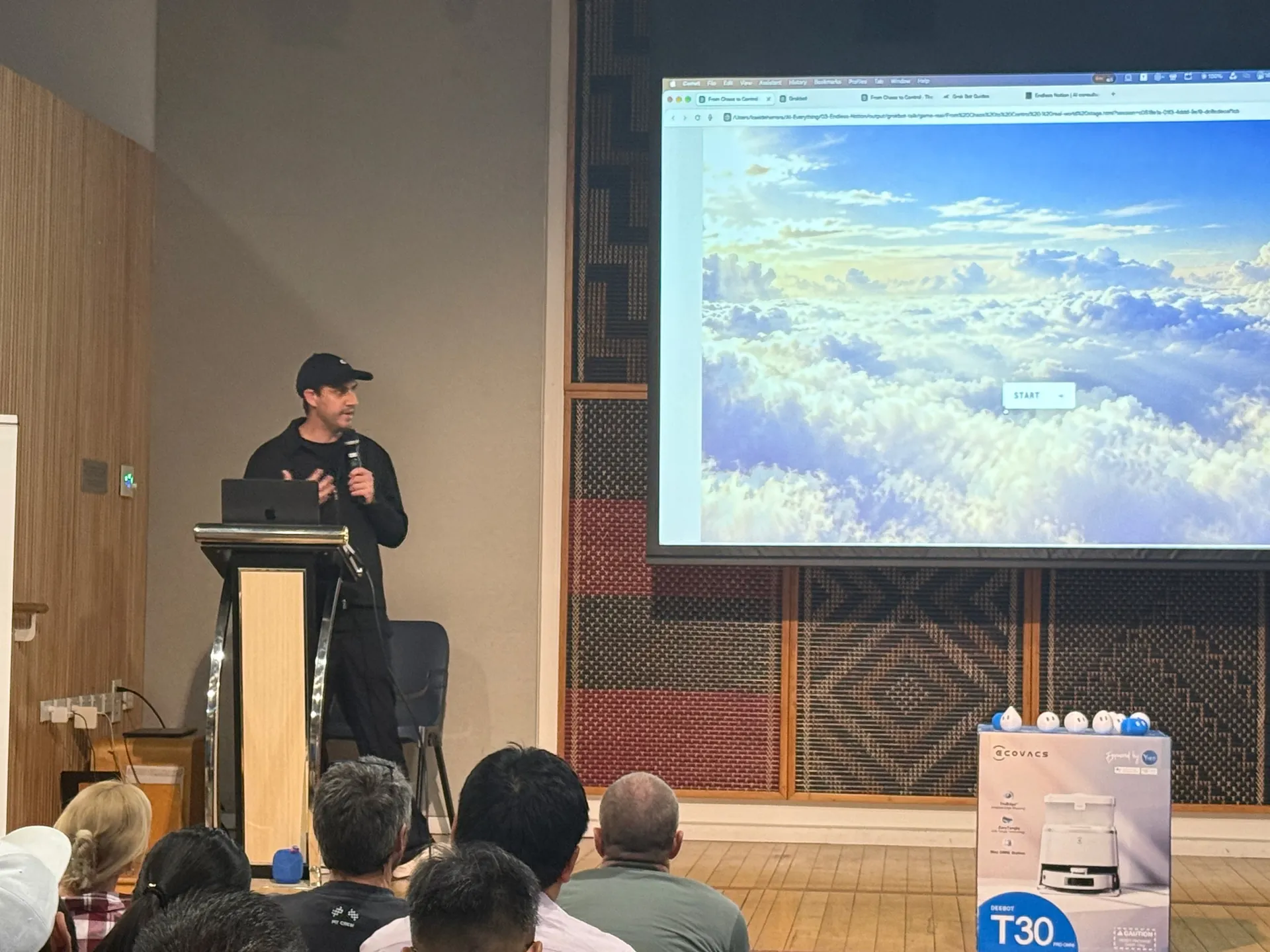 Speaker in a cap at the lectern with a clouds image on screen